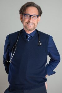 Male doctor in a blue sweater vest and shirt, wearing blue-framed glasses with a stethoscope around his neck, smiling at the camera.