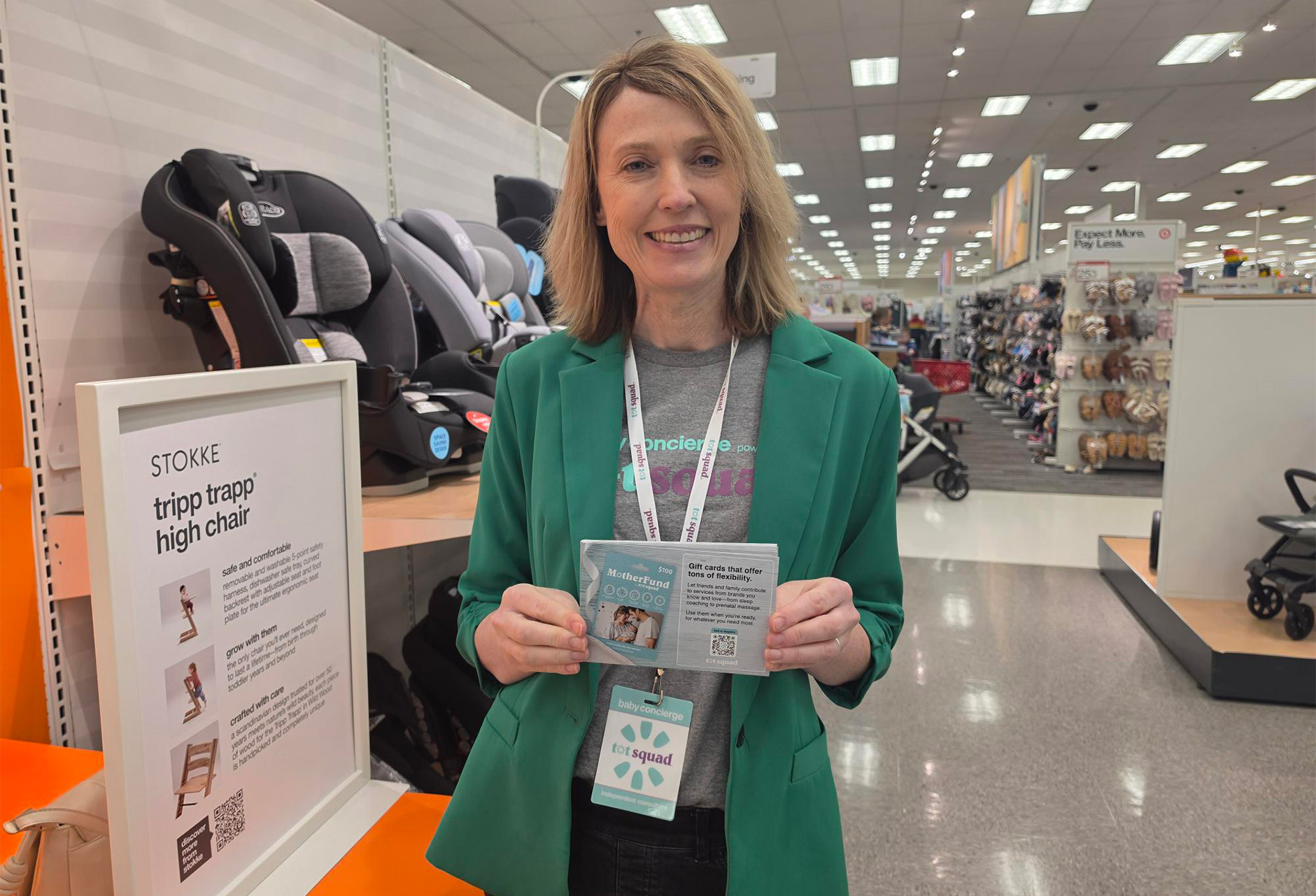 Smiling woman in a green blazer holding brochures at a store information desk with car seats on display behind her.
